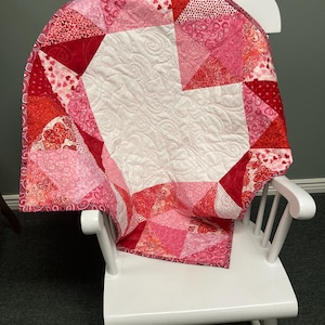 May include: A pink and red patchwork quilt with a white center and a heart-shaped design. The quilt is folded and placed on a white rocking chair.