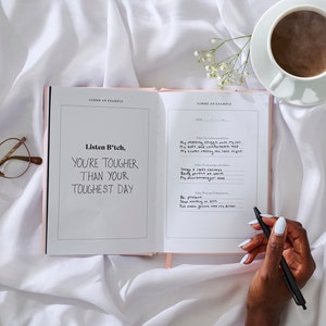 May include: Open journal with the quote "Listen B*tch, You're Tougher Than Your Toughest Day" written inside. A hand is holding a black pen over the open journal. A cup of coffee and a pair of glasses are on the white bedding.