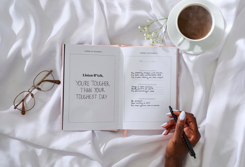 May include: Open journal with the quote "Listen B*tch, You're Tougher Than Your Toughest Day" written inside. A hand is holding a black pen over the open journal. A cup of coffee and a pair of glasses are on the white bedding.