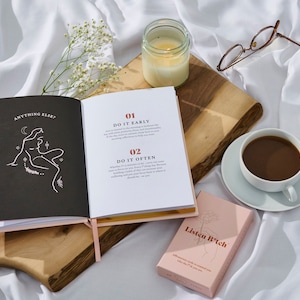 May include: Open book with the title "Anything Else?" and text that reads "01 Do It Early" and "02 Do It Often". A pink box with the text "Listen B*tch" is on a wooden surface with a white candle and a cup of coffee.