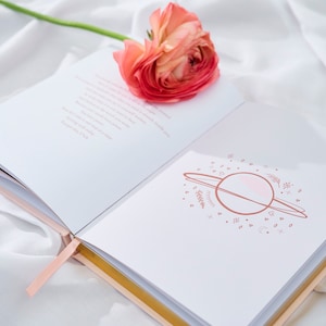 May include: An open journal with a pink flower on top. The journal has a pink ribbon bookmark and a gold-colored edge. The inside of the journal has a pink and white illustration of a planet with rings and stars.