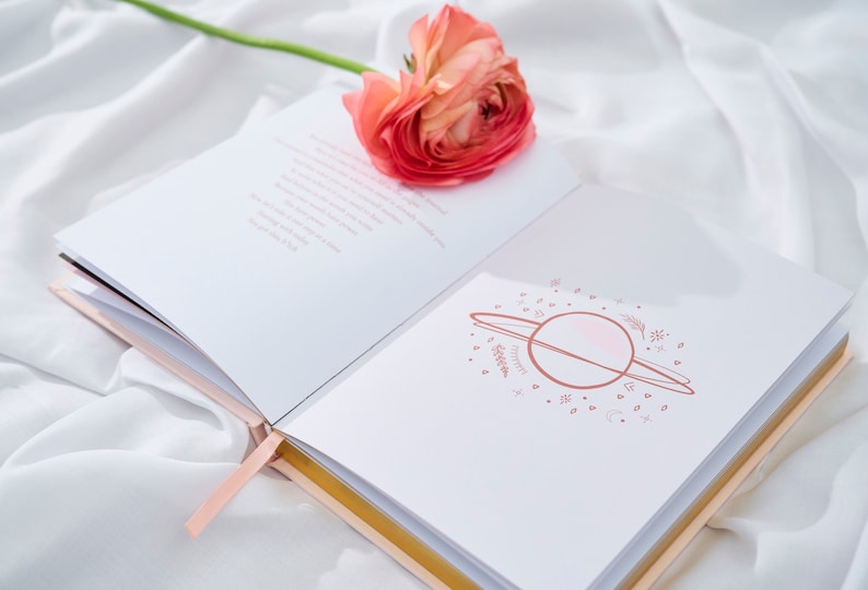 May include: An open journal with a pink flower on top. The journal has a pink ribbon bookmark and a gold-colored edge. The inside of the journal has a pink and white illustration of a planet with rings and stars.