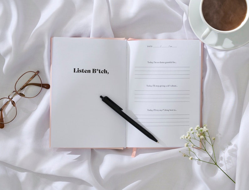 May include: Open pink journal with the text "Listen B*tch," on the left page. The right page has a date section and three lines with the text "Today, I'm so damn grateful for...", "Today, I'll stop giving a sh*t about...", and "Today, I'll try my f*cking best to..."