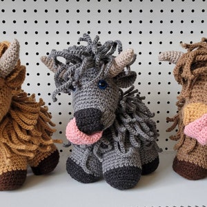May include: Three crocheted stuffed animals shaped like Highland cows. The cows are brown, gray, and brown with a pink nose. Each cow has a pink tongue sticking out.