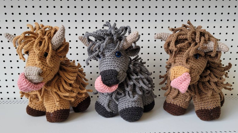 May include: Three crocheted stuffed animals shaped like Highland cows. The cows are brown, gray, and brown with a pink nose. Each cow has a pink tongue sticking out.