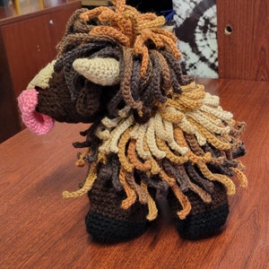 May include: A crocheted brown, tan, and orange fluffy animal with a pink nose. The animal has a long mane and a long tail.