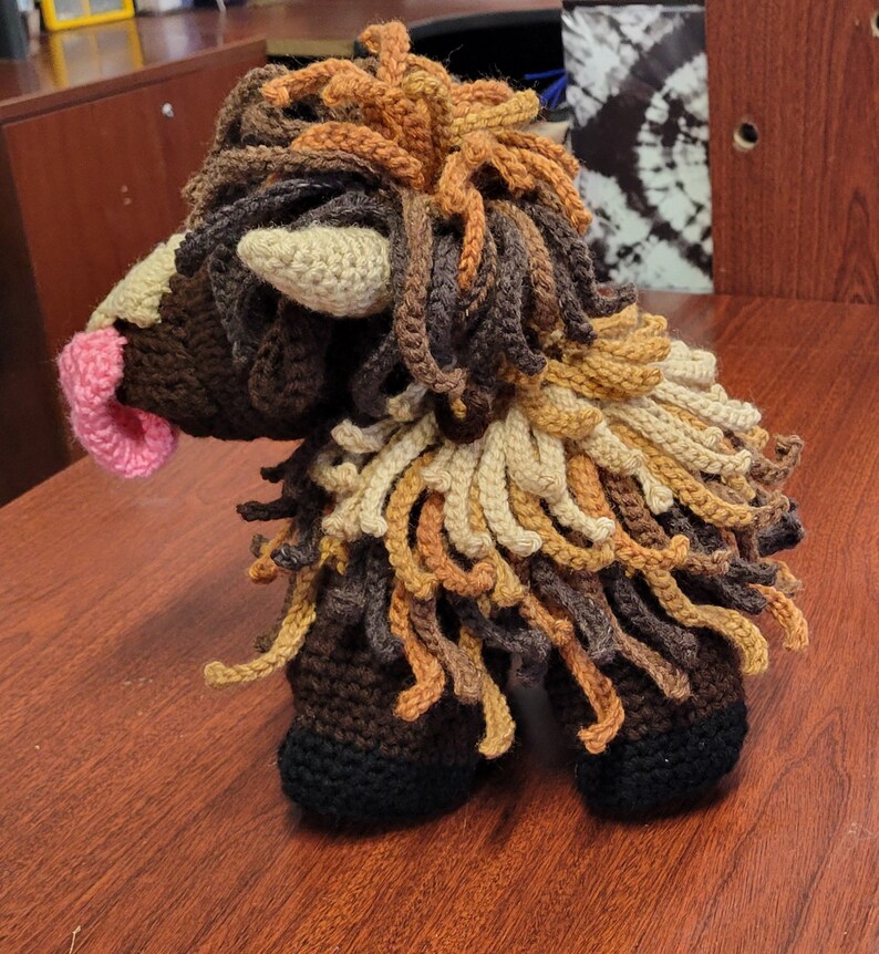 May include: A crocheted brown, tan, and orange fluffy animal with a pink nose. The animal has a long mane and a long tail.