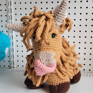 May include: A crocheted stuffed toy in the shape of a Highland cow. The cow is brown with a pink nose and green eyes. It has a long, shaggy mane and tail.