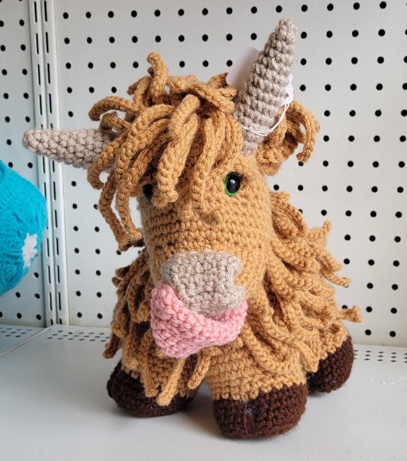 May include: A crocheted stuffed toy in the shape of a Highland cow. The cow is brown with a pink nose and green eyes. It has a long, shaggy mane and tail.