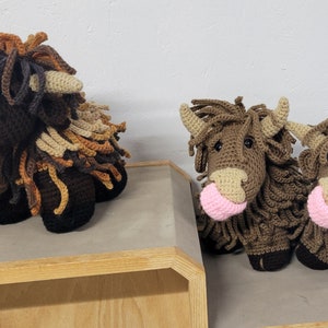 May include: Three crocheted stuffed highland cows with brown and tan fur. The cows have pink noses and are sitting on a wooden shelf.