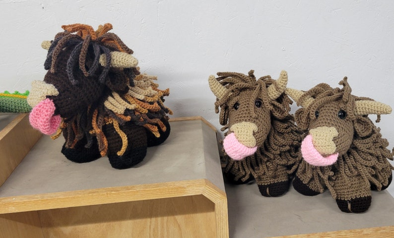 May include: Three crocheted stuffed highland cows with brown and tan fur. The cows have pink noses and are sitting on a wooden shelf.
