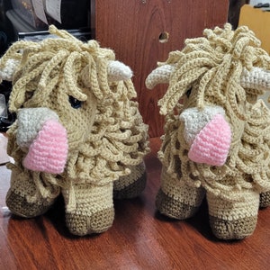May include: Two crocheted stuffed animals shaped like Highland cows. The cows are brown and white with pink noses and have long, shaggy fur.