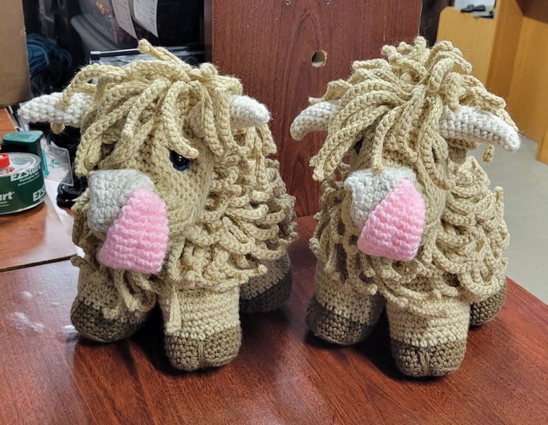 May include: Two crocheted stuffed animals shaped like Highland cows. The cows are brown and white with pink noses and have long, shaggy fur.