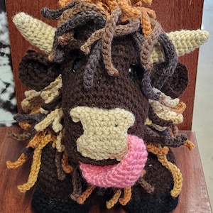 May include: A crocheted stuffed toy in the shape of a brown and white highland cow with a pink tongue sticking out. The cow has a long, shaggy mane and tail.
