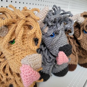 May include: Three crocheted stuffed animals shaped like Highland cows. The cows are brown, gray, and light brown with pink noses. The cows are all looking to the right.