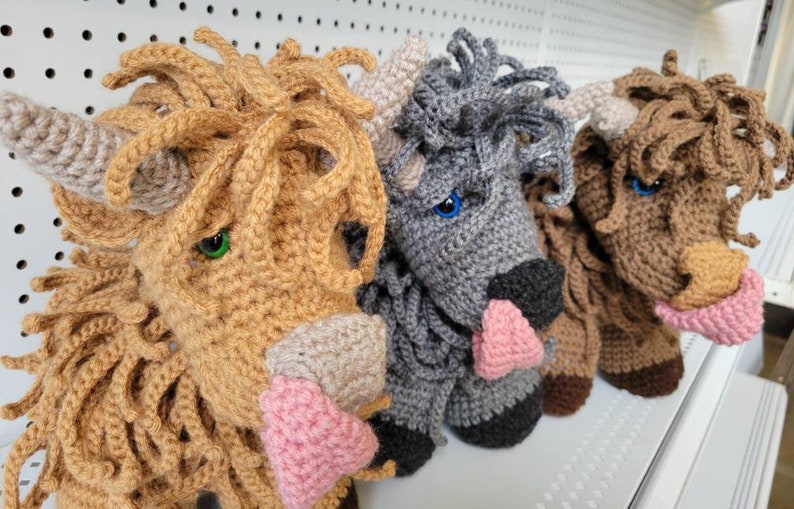 May include: Three crocheted stuffed animals shaped like Highland cows. The cows are brown, gray, and light brown with pink noses. The cows are all looking to the right.