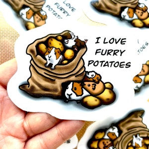 May include: A sticker featuring a brown hessian sack filled with guinea pigs and potatoes. The text on the sticker reads "I love furry potatoes".