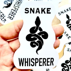 May include: White sticker with black text and a black snake design. The text reads "SNAKE WHISPERER".