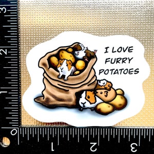 May include: A white sticker with a cartoon illustration of guinea pigs in a burlap sack with potatoes. The text "I love furry potatoes" is written in black.