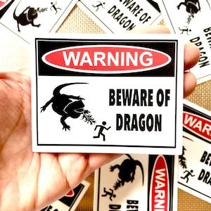 Puede incluir: Un cartel de advertencia blanco con bordes negros y rojos. El cartel dice "WARNING BEWARE OF DRAGON" en texto negro. Se muestra una silueta negra de un dragón con una persona corriendo en el cartel.