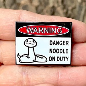 Peut inclure: Épinglette en émail noir et blanc avec un panneau d'avertissement qui indique "ATTENTION, DANGER NOODLE ON DUTY". Le panneau représente un serpent de dessin animé avec un visage souriant.