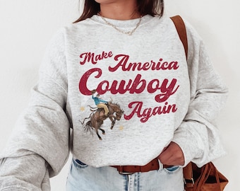 May include: A gray sweatshirt with the text "Make America Cowboy Again" in red lettering. A cartoon cowboy riding a bucking horse is printed on the front of the sweatshirt.