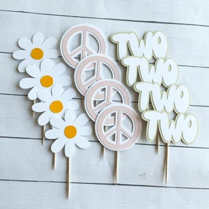 May include: A collection of decorative cake toppers featuring white daisies with orange centers, pink peace signs, and the word "TWO" in a playful font. The toppers are attached to wooden sticks.