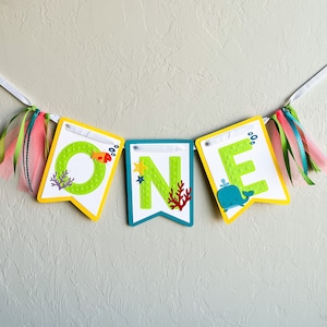 May include: A colorful banner with the word "ONE" spelled out in green letters with white dots. Each letter has a different ocean-themed design, including a fish, a starfish, coral, and a whale.
