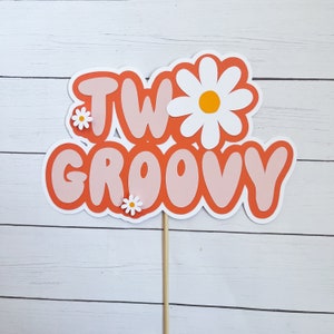 May include: Orange and pink cake topper with the text "TWO GROOVY" and white daisies.