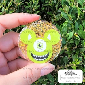 Puede incluir: Una agarradera de teléfono de resina verde y amarilla con una cara de monstruo de dibujos animados con un ojo y orejas de Mickey Mouse. El fondo está lleno de purpurina dorada.