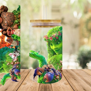 May include: A clear glass tumbler with a bamboo lid, featuring a colorful design of characters from a popular animated movie. The design includes images of a green-skinned character with a flower crown, and other characters. The tumbler sits on a wooden surface.