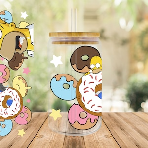 Op de afbeelding: Een transparante glazen beker met een bamboe deksel en een rietje. De beker heeft een cartoon afbeelding van een geel personage met een donut. De afbeelding bevat ook verschillende donuts in verschillende kleuren.