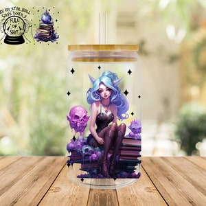 Puede incluir: Un vaso de vidrio transparente con tapa de bambú, con una ilustración de fantasía de una figura élfica de pelo azul sentada sobre libros, rodeada de calaveras y estrellas. El vaso tiene una pajita.