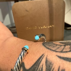 May include: A silver-toned twisted cable bracelet with gold accents and a turquoise stone. The bracelet is on a wrist with a tattoo. A brown box with the name "DAVID YURMAN" is in the background.