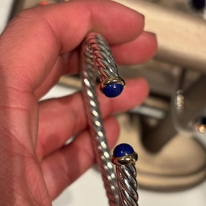 May include: A silver-toned, twisted metal bracelet with gold accents and two deep blue, round gemstones. The bracelet is open-ended and designed to slip over the wrist. The gemstones are set in gold bezels.