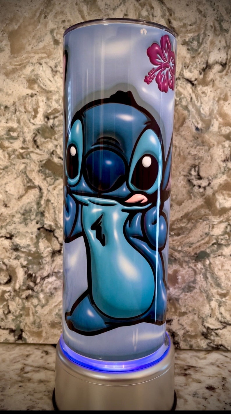 May include: A blue and white tumbler with a cartoon image of Stitch from Lilo & Stitch. The tumbler has a light blue base with a blue light.