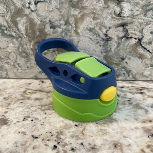May include: A green and blue water bottle lid with a carrying handle. The lid has a yellow button and a green flap.