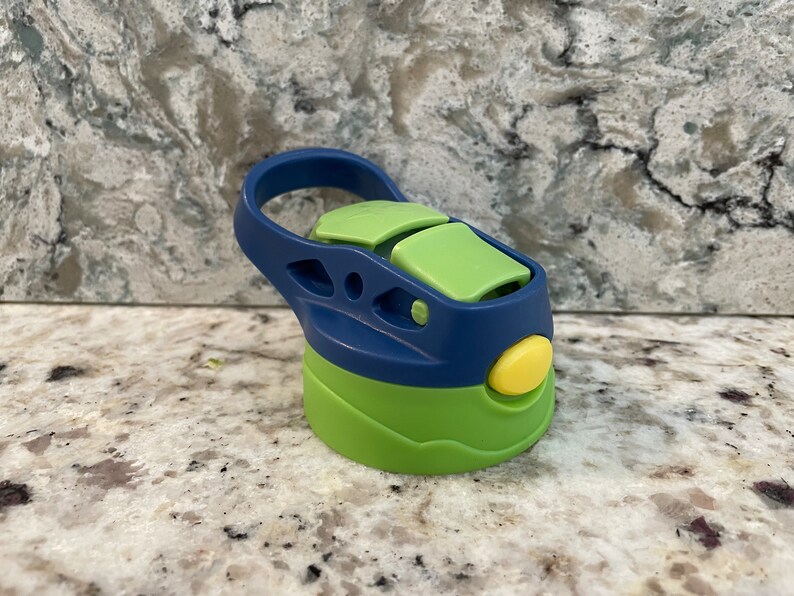 May include: A green and blue water bottle lid with a carrying handle. The lid has a yellow button and a green flap.
