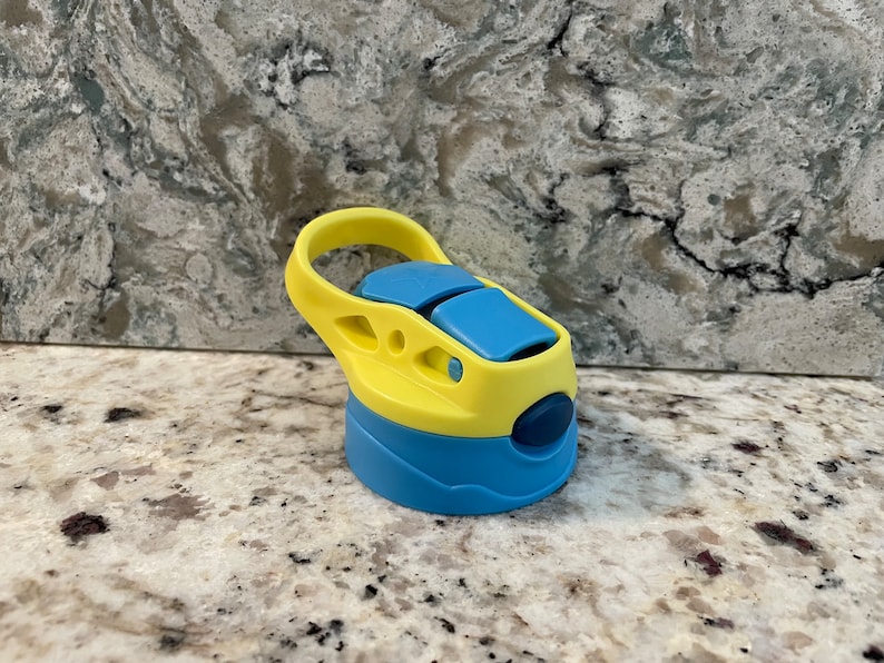 May include: A yellow and blue water bottle lid with a carrying handle. The lid has a blue button on the side.