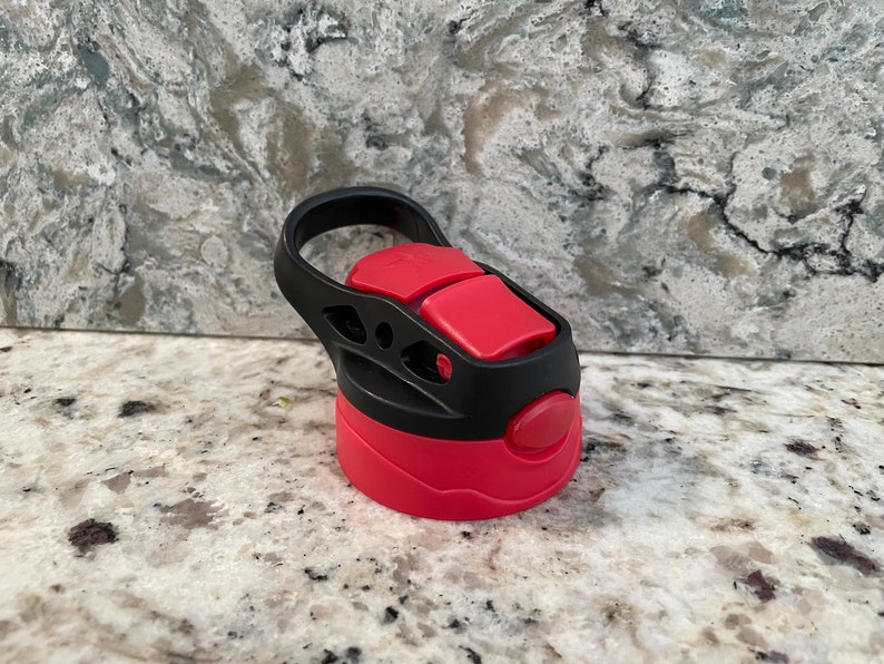 May include: A red and black water bottle lid with a carrying handle. The lid has a push button to open and close the spout.