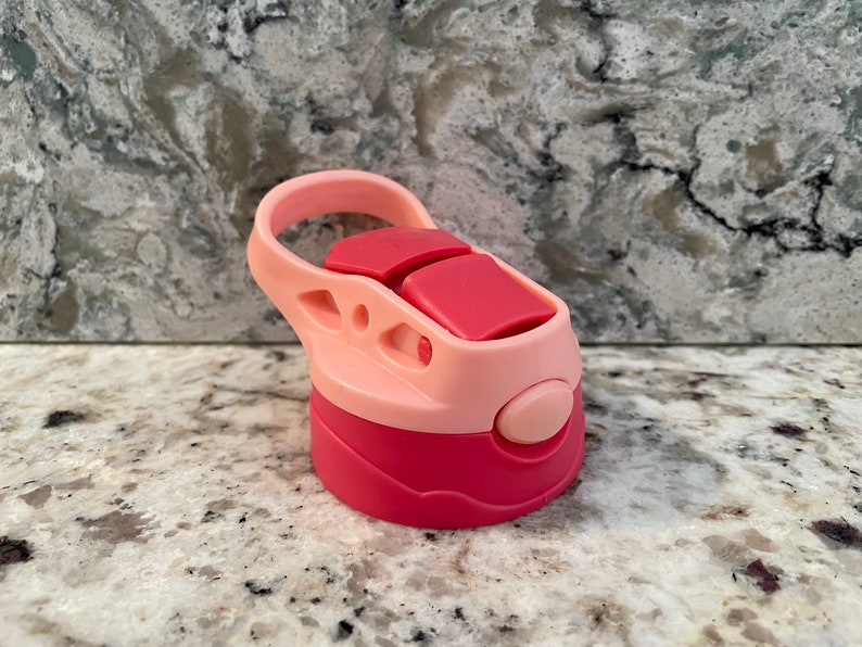 May include: Pink and red plastic water bottle lid with a carrying handle. The lid has a push button on the side.