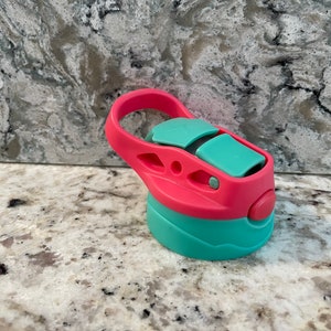 May include: A pink and teal water bottle lid with a carrying handle. The lid has a push button to open and close the spout.