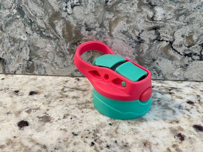 May include: A pink and teal water bottle lid with a carrying handle. The lid has a push button to open and close the spout.