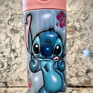 May include: A blue and white stainless steel water bottle with a cartoon image of Stitch from Lilo & Stitch. The bottle has a pink lid and a pink and purple base. The bottle is decorated with pink hibiscus flowers.