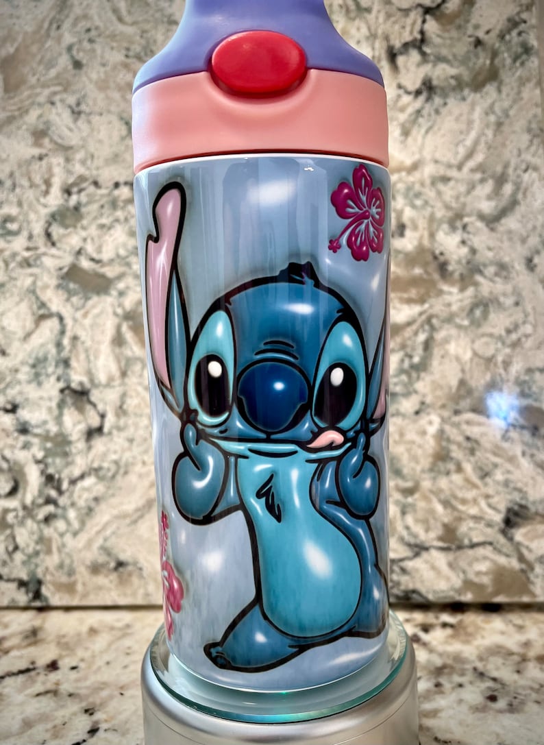 May include: A blue and white stainless steel water bottle with a cartoon image of Stitch from Lilo & Stitch. The bottle has a pink lid and a pink and purple base. The bottle is decorated with pink hibiscus flowers.