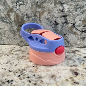 May include: A pink and purple water bottle lid with a carrying loop. The lid has a red button and a pink flap.