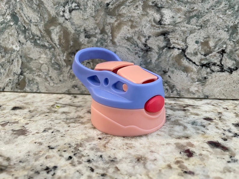 May include: A pink and purple water bottle lid with a carrying loop. The lid has a red button and a pink flap.