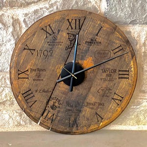 May include: A wooden clock face with Roman numerals and various bourbon brand names, including Woodford Reserve, Old Forester, Four Roses, Taylor, and Buffalo Trace. The clock hands are black with a gold accent.