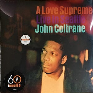 Może przedstawiać: Okładka albumu winylowego "A Love Supreme Live in Seattle" autorstwa Johna Coltrane'a. Okładka przedstawia portret Johna Coltrane'a i tytuł albumu w kolorze pomarańczowym, fioletowym i turkusowym. Zawiera logo Impulse! Records.