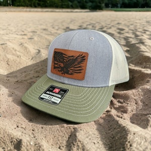 May include: A gray and white trucker hat with a green brim. The hat has a brown leather patch with an eagle and an American flag design. The patch says "EST 1970 RICHARDSON. ORIGINAL 112 TRUCKER ADJUSTABLE".